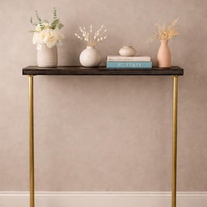 Puede incluir: Una consola de madera oscura con patas de metal dorado. La mesa está decorada con jarrones blancos con flores y plantas secas, y una pila de libros. El texto "IC STAST" es visible en un libro.