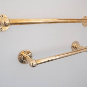 Op de afbeelding: Twee gouden metalen handdoekrekken gemonteerd aan een witte muur. De bovenste stang is langer dan de onderste stang.