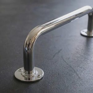 Puede incluir: Una barra de agarre cromada pulida con un diseño curvo. La barra está montada sobre dos bases circulares, cada una asegurada con tornillos. La superficie reflectante de la barra contrasta con el fondo oscuro.