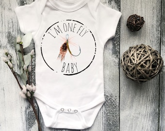 May include: White baby onesie with a black circle and the text "I'm one fly baby" inside the circle. A fishing fly and hook are inside the circle.