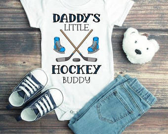 May include: White baby onesie with the text "Daddy's Little Hockey Buddy" and a graphic of two hockey sticks crossed with a puck and two blue ice skates.