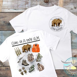 Puede incluir: Camiseta blanca con un gr&aacute;fico de un oso marr&oacute;n y el texto "Going on a Bear Hunt". La parte posterior de la camiseta presenta un oso, un chaleco, binoculares, un rifle, botas, municiones y un abrigo.