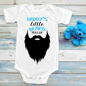 May include: White baby onesie with the text "Daddy's little beard puller" in blue and black. A black silhouette of a beard is below the text.