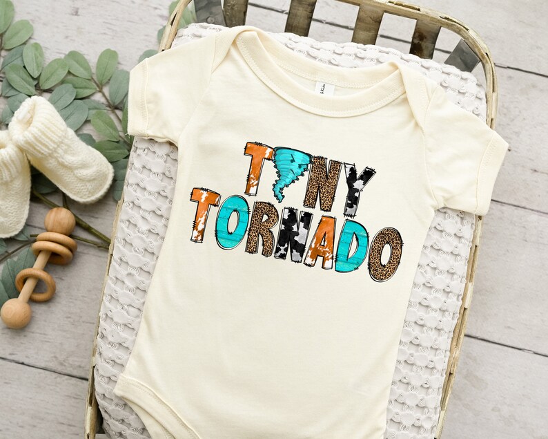 Op de afbeelding: Een witte baby romper met de tekst "Tiny Tornado" erop gedrukt in kleurrijke letters met dierenprintpatronen. De letters zijn zwart omrand en het woord "Tornado" heeft een blauwe en witte tornado-afbeelding.