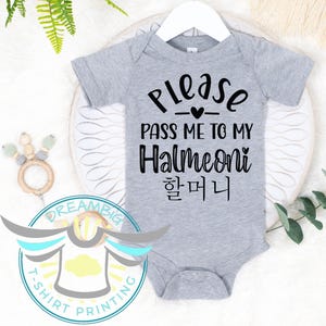 May include: A gray baby onesie with the text "Please pass me to my Halmeoni" in black. The onesie is hanging on a white hanger. A wooden teething ring and a logo are visible in the background.