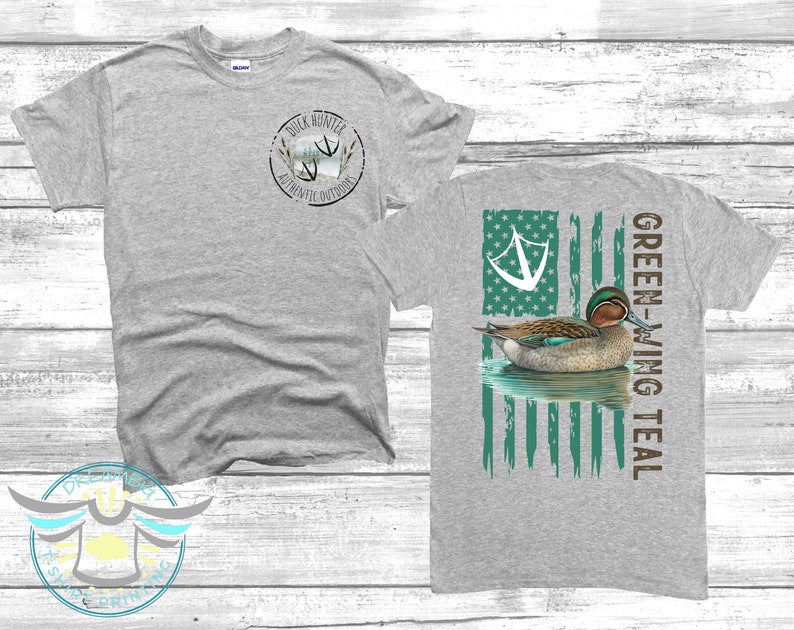 May include: Gray t-shirt with a green-winged teal duck on a distressed American flag design on the back. The front of the shirt has a small circle logo with the text "Duck Hunter Authentic Outdoors".