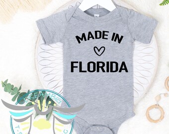 May include: A gray baby onesie with the text "MADE IN" above a heart symbol and the word "FLORIDA" in bold black letters. The onesie is hanging on a white hanger.