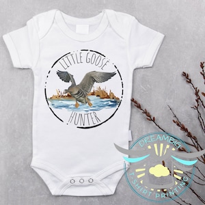 May include: White baby onesie with a black and white circle design. The circle has the text "Little Goose Hunter" and a watercolor illustration of a goose flying over a lake.