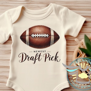 May include: A white baby bodysuit with a brown football graphic and the text "Newest Draft Pick".