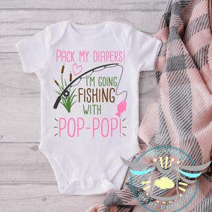 May include: White baby onesie with the text "PACK MY DIAPERS! I'M GOING FISHING WITH POP-POP!" in pink and brown. The design includes a fishing rod, a pink fish, and green cattails. A cute baby outfit.