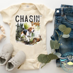 May include: A cream-colored baby onesie with the text "CHASIN' tails" in black and brown lettering. A watercolor illustration of a beagle dog chasing a rabbit is printed on the onesie.