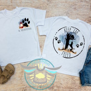 May include: Two white t-shirts with different designs. The first shirt has a red lantern and paw print with the text "Ol' Red Outfitters". The second shirt has a black silhouette of a dog in a tree with the text "Keep 'em treed".