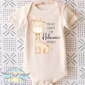 May include: Cream-colored baby onesie with a giraffe graphic. The giraffe is light brown with darker brown spots. Text on the onesie reads "YOU BET GIRAFFE I'M Halmeoni's FAVORITE". The onesie has short sleeves and snap closures.