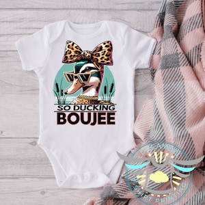 May include: White baby bodysuit with a graphic of a duck wearing sunglasses and a leopard print bow. The duck is set against a teal background with cattails. The text "SO DUCKING BOIJEE" is below the duck.