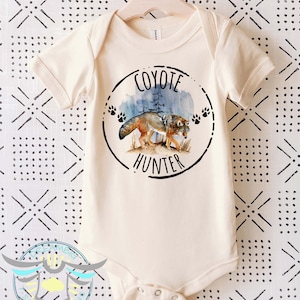 May include: A cream-colored baby onesie with a black and white graphic of a coyote in a forest setting. The graphic includes the text "Coyote Hunter".