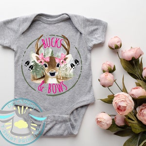 May include: A gray baby onesie with a white circle design featuring a cartoon deer with pink bows and the text "Bucks & Bows".
