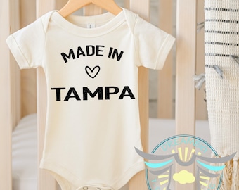 May include: Cream-colored baby onesie with the text "MADE IN" above a heart symbol and the word "TAMPA" in bold black letters. The onesie hangs on a wooden hanger against a crib.