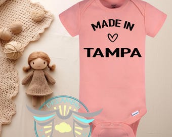 May include: A light pink baby onesie with the text "MADE IN TAMPA" in black. A small heart is between the words. A doll and other decorative items are in the background.