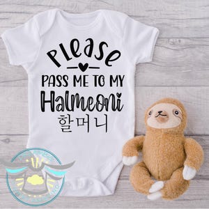 May include: White baby onesie with black text that reads "Please pass me to my Halmeoni 할머니". A brown sloth plush toy is in the background.