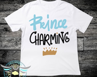 Puede incluir: Camiseta blanca con el texto "Prince Charming" en letras negras y azules. Una corona dorada se encuentra debajo del texto.