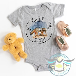 May include: A gray baby onesie with a circular graphic featuring a coyote illustration and the text "COYOTE HUNTER". The design includes paw prints and a forest scene. A plush teddy bear and a pair of rose gold baby shoes are also visible.