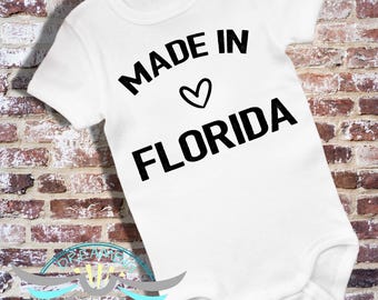 May include: White baby bodysuit with the text "MADE IN FLORIDA" in black, with a heart symbol. The bodysuit is made of soft material and is designed for infants. The background is a brick wall.