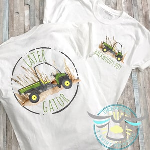 May include: Two white t-shirts with a green and black utility vehicle design. One shirt says "Later Gator" with a circle around the vehicle. The other shirt says "Backwoods Boy" with the vehicle in the center.
