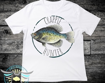 May include: A white short-sleeved t-shirt featuring a central graphic of a speckled fish inside a dark teal circular border. The text CRAPPIE HUNTER is printed in black capital letters above and below the fish illustration on the front of the shirt.
