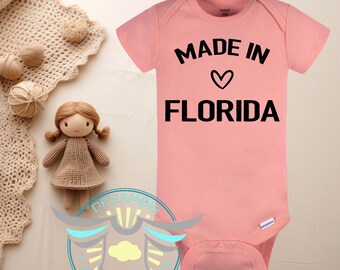 May include: A light pink baby onesie with the text "MADE IN FLORIDA" in black. A heart symbol is between the words "IN" and "FLORIDA". The onesie is on a light beige surface with a doll and decorative items.