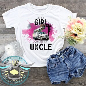 May include: White t-shirt with the text "JUST A GIRL WHO LOVES HER UNCLE" and a graphic of a utility truck. The design includes pink and gray watercolor-style splashes. Paired with denim shorts.