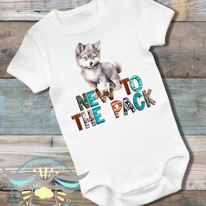 May include: White baby onesie with a gray wolf pup illustration and the text "NEW TO THE PACK" in colorful letters with a tribal pattern.