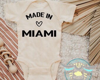 May include: Cream-colored baby onesie with the text "MADE IN" above a heart symbol and "MIAMI" in bold black letters. The onesie is short-sleeved and has a simple, classic design, perfect for a newborn.