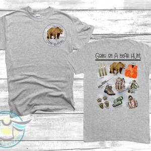 Puede incluir: Dos camisetas grises con tem&aacute;tica de caza de osos. La parte delantera de una camiseta tiene un oso marr&oacute;n con el texto "Gone Hunting". La parte trasera de la otra camiseta tiene el texto "Going on a Bear Hunt" e ilustraciones de equipo de caza, incluyendo un oso, un rifle, botas y un chaleco de caza.