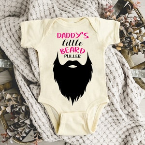 May include: A cream-colored baby onesie with the text "Daddy's little beard puller" in black and pink lettering. A black silhouette of a beard is below the text.