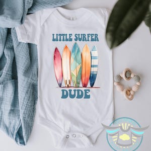 May include: White baby bodysuit with the text "LITTLE SURFER DUDE" above a row of five colorful surfboards. The surfboards are in shades of pink, orange, green, and blue. A baby toy is in the upper right corner.
