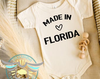 May include: Cream-colored baby bodysuit with the text "MADE IN FLORIDA" in black, with a heart symbol. A wooden rattle is next to the bodysuit. The bodysuit is on a beige surface.