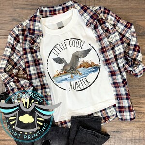 May include: White t-shirt with a circular design featuring a goose in flight and the text "Little Goose Hunter". The shirt is laid out on a plaid flannel shirt and black jeans.
