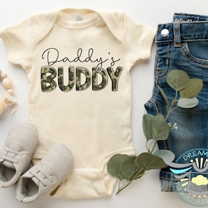 May include: A cream-colored baby onesie with the text "Daddy's Buddy" in black lettering with a camouflage pattern. The onesie is next to a pair of gray baby shoes and a pair of blue jeans.