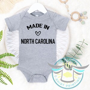 May include: A grey baby bodysuit featuring the text "MADE IN" above a heart symbol and "NORTH CAROLINA" in black. The bodysuit is suspended from a white hanger.