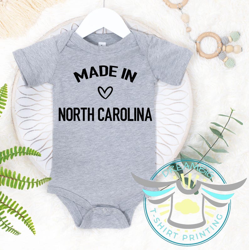 May include: A grey baby bodysuit featuring the text "MADE IN" above a heart symbol and "NORTH CAROLINA" in black. The bodysuit is suspended from a white hanger.