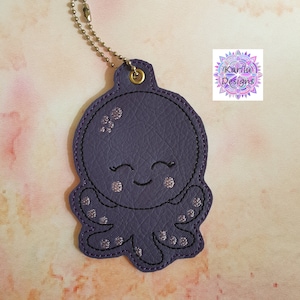 May include: Purple leather octopus keychain with a smiling face and silver glitter accents. The keychain has a metal ring and chain.