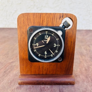 May include: A vintage black and silver 8-day clock with white numerals, set in a wooden stand. The clock face displays the numbers 12, 3, 6, and 9, with smaller markings for minutes. The wooden stand has a rectangular shape.