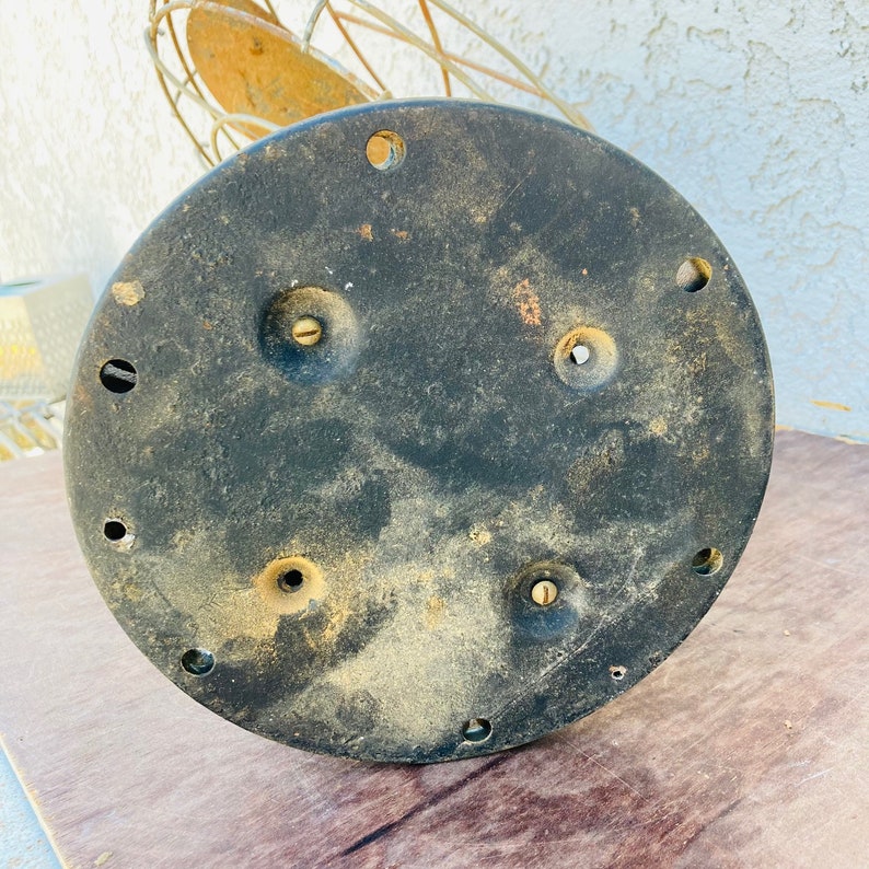 Pu&ograve; includere: Il lato inferiore di una base di ventilatore vintage in metallo. La base &egrave; nera e ha diversi fori per viti.