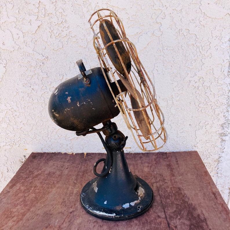 Pu&ograve; includere: Un ventilatore vintage in metallo nero con una gabbia di filo arrugginito e due pale. Il ventilatore &egrave; su un tavolo di legno.