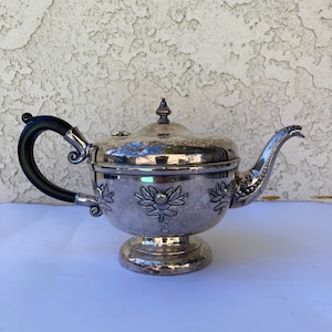 May include: Silver teapot with a black handle and floral design. The teapot has a round body and a lid with a finial.
