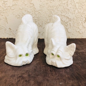 May include: Two white ceramic dog figurines with green eyes. The dogs are in a crouching position with their heads down, as if they are sniffing the ground.