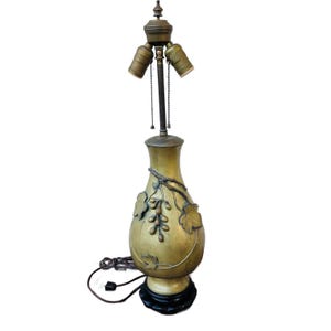 May include: A gold table lamp with a vase-shaped base featuring an embossed grapevine design. The lamp has a black base and two light bulbs.