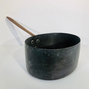 May include: A vintage copper saucepan with a wooden handle. The saucepan has a dark patina and four small rivets around the rim.