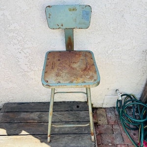 May include: An antique metal chair with a square seat and backrest. The chair has a distressed, weathered appearance with a light blue finish and rust-colored seat. The legs are slender and the chair is set on a wooden surface.