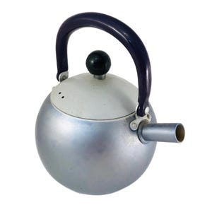 May include: A silver-colored, spherical kettle with a dark blue handle and a black knob on the lid. The spout is cylindrical and extends from the kettle's side. The lid is white. A classic kitchen appliance.